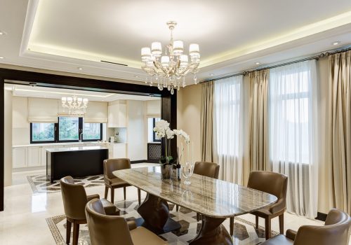Classic interior of dining room in brown, white, beige colors, with crystal chandelier in the center of ceiling. Wooden table and many chairs around.View of other room with kitchen bar.